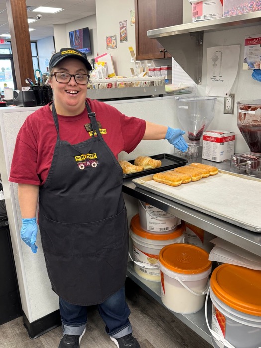A Sweet Street team member in gloves and apron smiling beside trays of fresh bakes